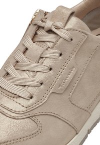 Beige athletic shoe featuring a suede texture, metallic accents, flat laces, and a stitched design, with a white rubber sole.
