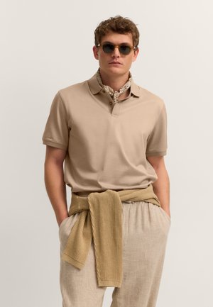 Mann trägt beiges kurzärmeliges Polo, hellen Schal um den Hals, Sonnenbrille, beigen Pullover um die Taille gebunden und lockere Leinenhose mit den Händen in den Taschen.