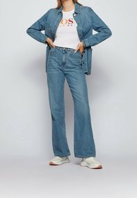 Chaqueta de mezclilla en azul claro, abotonada, con dos bolsillos en el pecho, combinada con una camiseta blanca con letras multicolores y jeans azules de campana.