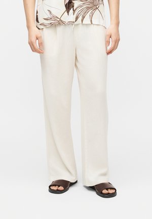 Person trägt lockere weiße Hose, braune Sandalen mit offenem Zehenbereich und ein beiges Hemd mit dunklen Palmenblattmustern, steht vor einem einfarbigen Hintergrund.