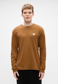 Καφέ μακρυ�μάνικο πουκάμισο με υφή, στρογγυλή λαιμόκοψη και λογότυπο στο στήθος. Χαλαρή εφαρμογή με ραμμένα μανίκια.