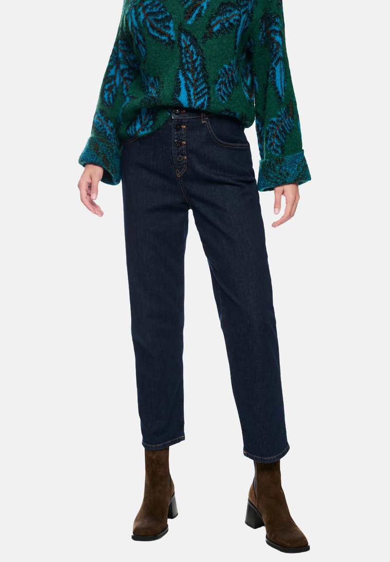 Dark blue denim jeans feature a high waist, five-button closure, and tapered ankle. Paired with a green sweater with blue leaf patterns.