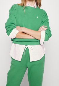 Zelená mikina z měkké tkaniny s žebrovanými manžetami, navrstvená přes světle růžovou košilovou košili. K tomu ladící zelené tepláky se šňůrkou.