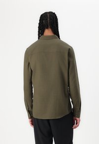 Camisa de color verde oliva con botones, hecha de un tejido texturizado, que presenta un cuello y mangas largas con puños abotonados. Diseño liso en la parte trasera.