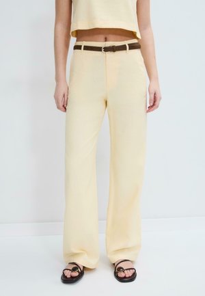 Kvinde iført beige bukser med vide ben og brun bælte, kombineret med sorte sandaler med runde spænder, stående mod en hvid baggrund.