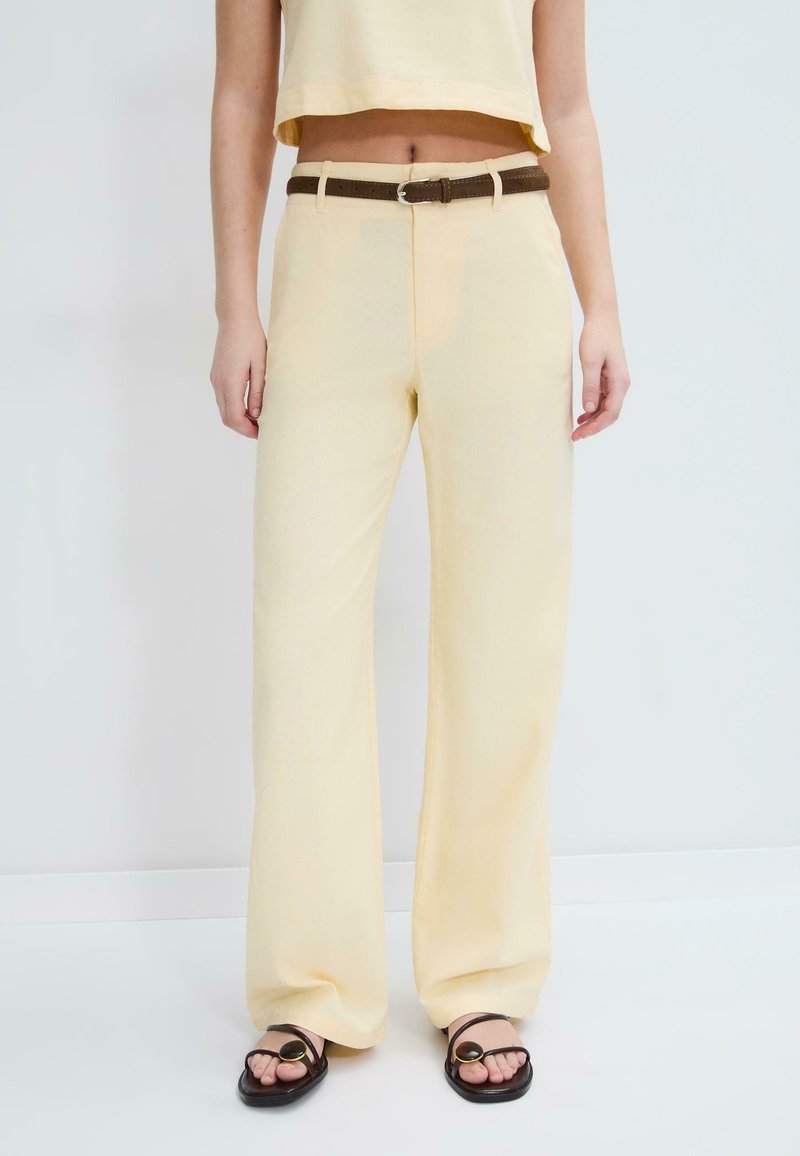 Femme portant un pantalon beige à jambes larges avec une ceinture marron, associé à des sandales noires avec des détails de boucle circulaire, debout sur un fond blanc.