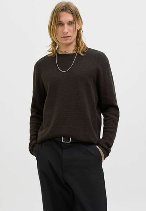 Dunkelbrauner gestrickter Pullover mit lockerer Passform, Rundhalsausschnitt und langen Ärmeln, kombiniert mit schwarzen Hosen und einer silbernen Halskette.