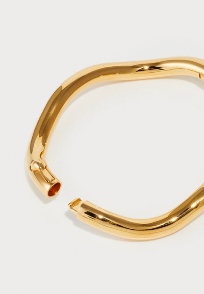 Brățară din metal auriu cu un design neted și curbat. Remarcabilă prin finisajul său strălucitor și capetele deschise, creând o apariție minimalistă.