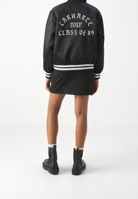 Schwarze Bomberjacke mit weißem Aufdruck "Carhartt WIP Class of 89", gestreiften Bündchen und Saum. Getragen über einem schwarzen Rock und kombiniert mit schwarzen Stiefeln.