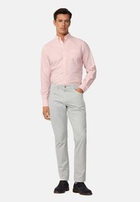 Camicia button-down rosa chiaro con colletto e tasca, abbinata a pantaloni grigi chiari. Il modello indossa scarpe scure e si trova davanti a uno sfondo bianco.