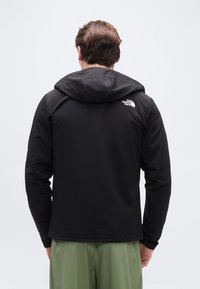 Homme portant une veste à capuche noire North Face et un pantalon vert, vu de dos sur un fond clair uni.