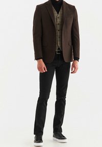 Blazer marrone sopra un gilet a quadri, abbinato a pantaloni neri e sneakers nere. Il blazer presenta una tasca sul petto e un design elegante.