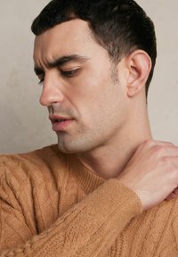 Sweter dzianinowy w ciepłym odcieniu brązu, z teksturowanymi wzorami w warkocze i ściągaczami przy mankietach. Zbliżenie na profil męskiego modela oraz jego rękę na ramieniu.