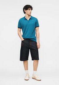 Jeune homme aux cheveux bouclés portant un polo turquoise, un short noir jusqu'aux genoux, des chaussettes blanches et des baskets beige, debout avec une main dans la poche.