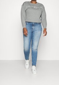 Sudadera gris con logo, jeans de mezclilla azules y zapatillas blancas. El conjunto presenta un corte relajado y elementos de diseño casual.