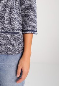 Blau-weißer, strukturiert gewebter Oberteil mit Dreiviertelärmeln, ausgefransten Bündchen und Rundhalsausschnitt, kombiniert mit hellblauer Jeans.