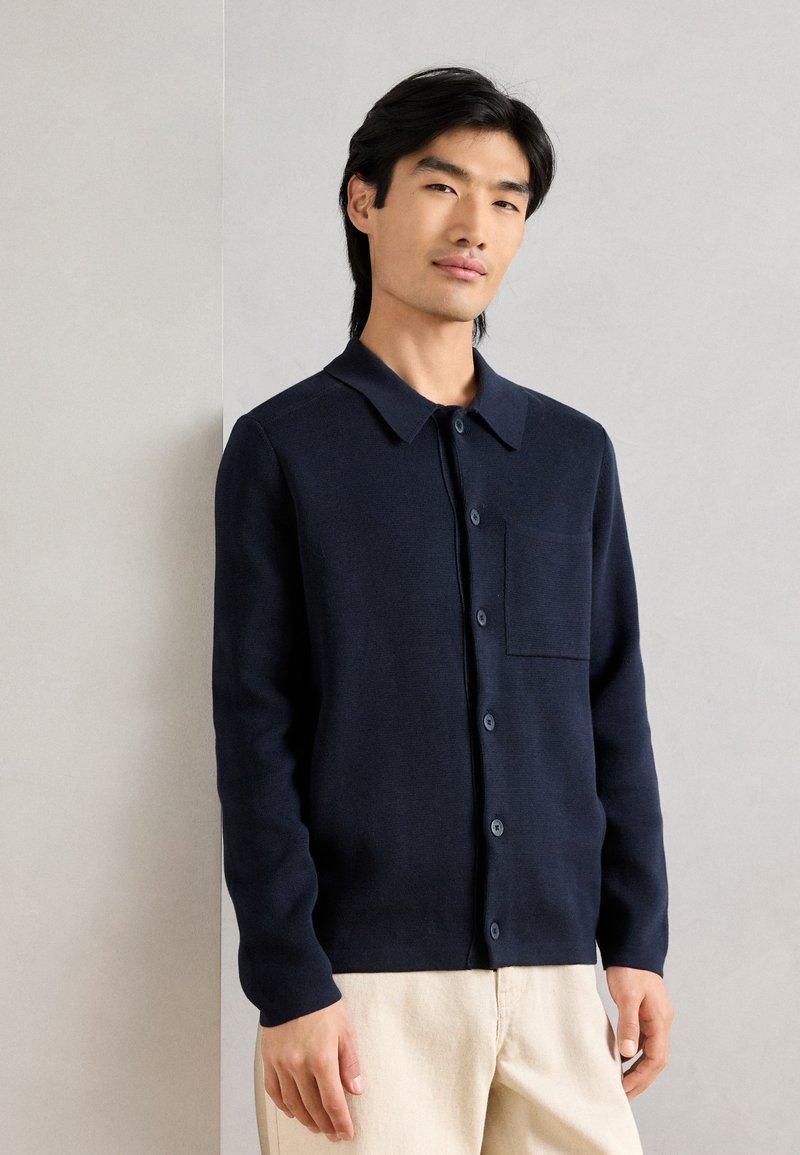 Μπλε πλέκτο πουλόβερ με κουμπιά μπροστά, γιακά και τσέπη στο στήθος. Έχει μακριά μανίκια και στενή εφαρμογή, κατασκευασμένο από μαλακό ύφασμα.