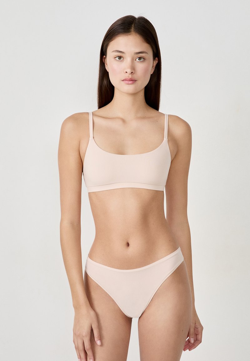 Junge Frau mit langen braunen Haaren, die ein leicht beige Unterwäsche-Set trägt, steht vor einem schlichten weißen Hintergrund und blickt nach vorne.