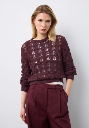 Mujer con cabello rubio vistiendo un suéter de punto color púrpura oscuro sobre una camiseta blanca y pantalones de talle alto a juego en púrpura oscuro.