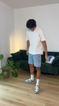 Νέος άνδρας με λευκό μπλουζάκι και τζιν σορτς κοιτάζει προς τα κάτω, στέκεται σε ξύλινο πάτωμα κοντά σε σκούρο πράσινο καναπέ με μαξιλάρια και φυτά εσωτερικού χώρου.