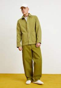 Chaqueta de trabajo verde oliva con dos bolsillos frontales y costuras blancas, combinada con pantalones holgados a juego y zapatillas blancas. Fondo amarillo.