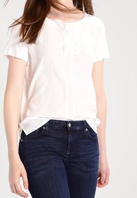 Blusa bianca a maniche corte con scollatura tonda e dettagli di pieghe verticali, abbinata a jeans in denim blu scuro. Tessuto liscio e leggero.