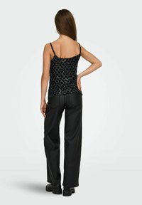 Femme de dos portant un haut à fines bretelles noir à motifs, un pantalon noir et des chaussures grosses noires sur un fond uni.