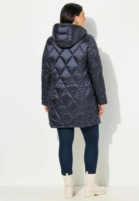 Maritiem gewatteerd pufferjack met capuchon, met een ruitpatroon. Het materiaal lijkt glanzend en soepel. Draagt over donkere jeans en lichte laarzen.