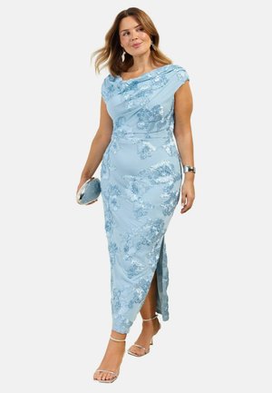 Kobieta w jasnoniebieskiej sukience z kwiatowym haftem i dekoltem odsłaniającym ramiona, trzyma srebrną kopertówkę i nosi srebrne szpilki.