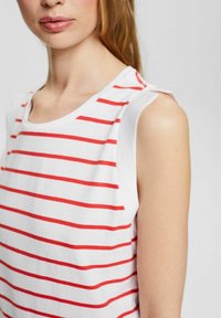 Top blanco sin mangas con rayas horizontales rojas, con lazos en los hombros. Hecho de una tela suave con un ajuste relajado, exhibiendo un diseño simple.