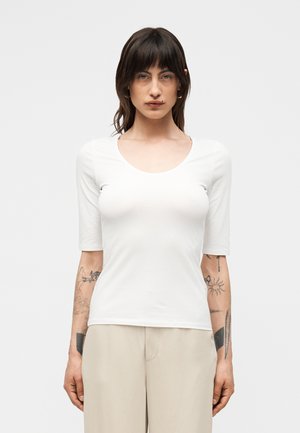 Ženska z rjavimi lasmi in tetovažami na rokah, oblečena v belo majico z okroglim ovratnikom in rokavi do komolcev ter bež hlače, stoji obrnjena naprej.