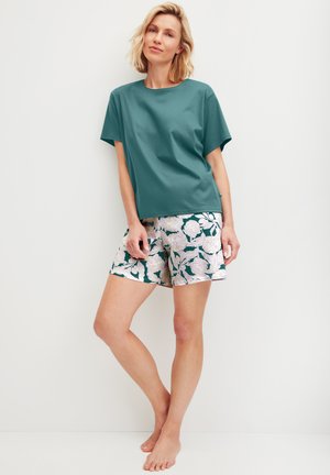 Blonde Frau steht barfuß, trägt ein türkisfarbenes kurzärmeliges Shirt und Shorts mit Blumenmuster vor einem einfarbigen weißen Hintergrund.