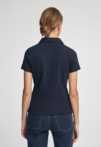 Marineblaues kurzärmliges Polohemd mit Kragen, glatter Textur und tailliertem Design, kombiniert mit dunklen Jeans.