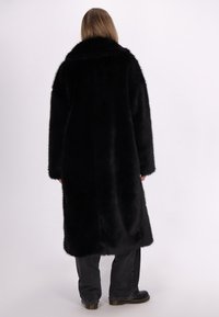 Sort faux fur jakke, lang og oversized, med bred krave og løse ærmer. Glat tekstur og strømlinet silhuet. Sættet ledsages af mørke bukser.