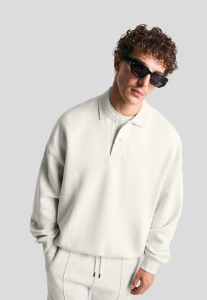 Lys grå pullover med krave, der har en afslappet pasform og blød tekstur. Kombineret med matchende lys grå sweatpants. Sorte solbriller bæres.