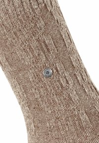 Beżowa, dziana skarpetka z teksturowanym wzorem, z wypukłymi geometrycznymi kształtami i metalowym akcentem z logo. Wykonana głównie z miękkiego i ciepłego materiału.