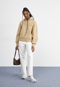 Beige hoodie with a front pocket and embroidered logo, paired with white straight-leg jeans, animal print bag, and gray sneakers.