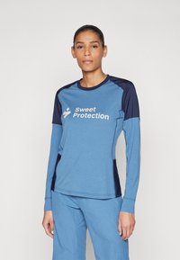 Gaišā zilā krāsā garām piedurknēm topiņš ar tumši zilu akcentu, ar apaļu dekoltē un "Sweet Protection" logo priekšpusē. Mīksta auduma, sportiska piegriezuma.