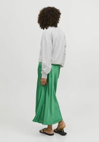 Pelēks treniņjaka, zaļš satīna maksi svārki ar plūstošu formu, apvienots ar melnā sandales. Modelis skatīts no aizmugures.