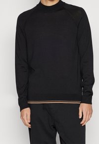 Czarny sweter z długim rękawem, z wysokim kołnierzem, ściągaczami przy rękawach oraz poziomym brązowym paskiem u dołu. Wykonany z gładkiej tkaniny.