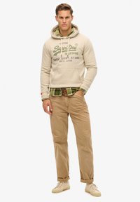 Felpa beige con scritte verdi sovrapposta a una camicia a quadri verdi con colletto, abbinata a pantaloni marrone chiaro e sneakers crema.