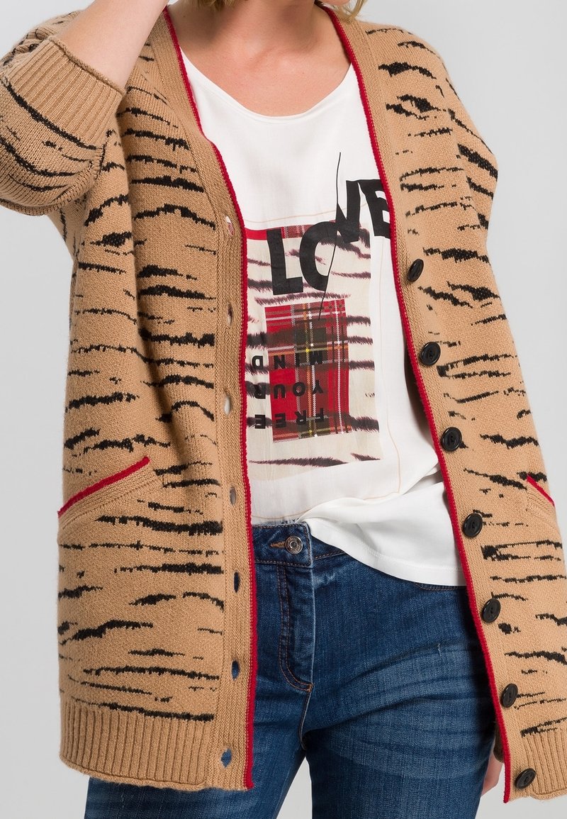Femme portant un jean bleu, un t-shirt blanc à motif graphique rouge et noir, et un cardigan beige boutonné avec des rayures noires et une bordure rouge.