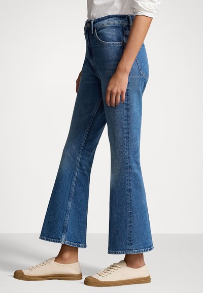 Uitgebrede blauwe denim jeans met een hoge taille, vervaagde textuur en onafgewerkte zoom. Gecombineerd met lichtbruine canvas sneakers met bruine zolen.