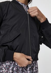 Černá bomber bunda s předním zipem, žebrovanými manžetami a náprsenkovou kapsou. Hladká textura, lehký materiál, stylizováno s květinovými šaty pod nohavicí.