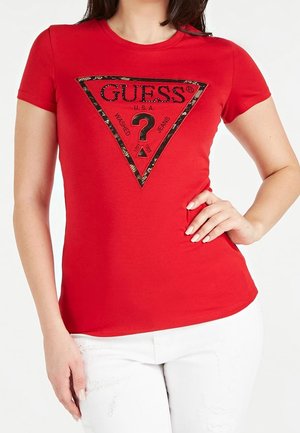 Kvinde iført en tætsiddende rød t-shirt med et stort sort og guld Guess-logo og hvide bukser, hendes højre hånd på hoften.