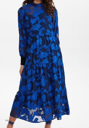 Mujer con vestido azul marino de manga larga con grandes estampados florales en azul brillante y falda de volantes, combinada con zapatos azules marino con cordones.