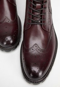 Burgundské kožené boty s perforovanými brogue detaily, kulatou špičkou a černou gumovou podrážkou. Mají ploché tkaničky a dekorativní švy.