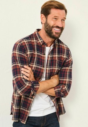 Flanellhemd mit einem rot, marineblau und beige karierter Muster, Knopfkragen und hochgekrempelten Ärmeln, getragen über einem weißen T-Shirt.