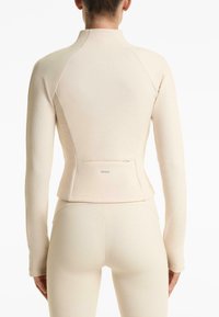Maglietta a maniche lunghe color beige chiaro con tessuto testurizzato, design aderente e tasca con zip sulla schiena. Presenta maniche raglan e un colletto alto.