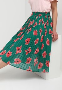 Gonna plissettata verde con motivo floreale rosso, vita elasticizzata e tessuto leggero. Accenti di texture aggiungono profondità al design.
